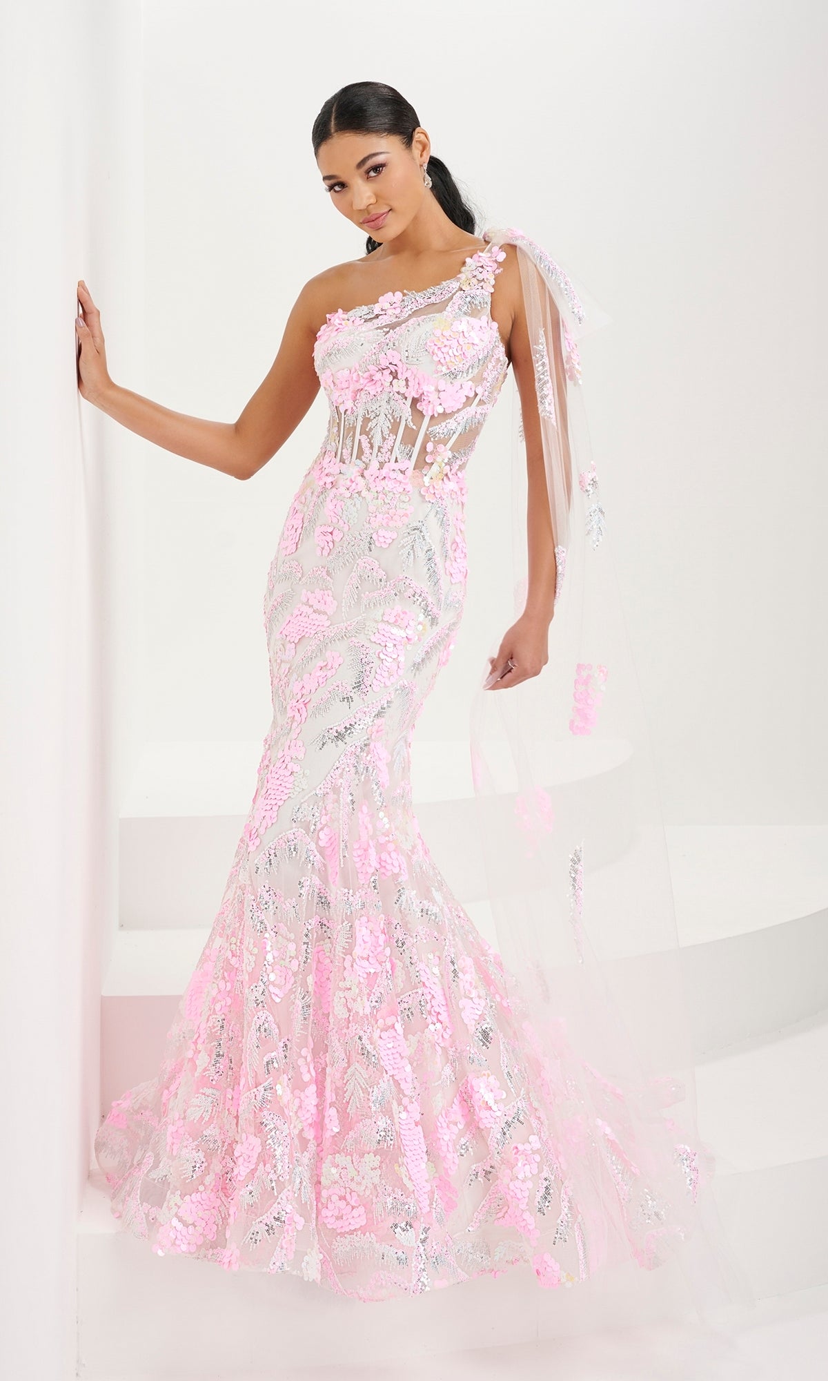 A woman poses in the Tiffany One-Shoulder Sequin Sheer Prom Dress 16082, featuring a fitted mermaid silhouette, sheer corset bodice, sequin floral embroidery, and a tulle bow on one shoulder against a white background.